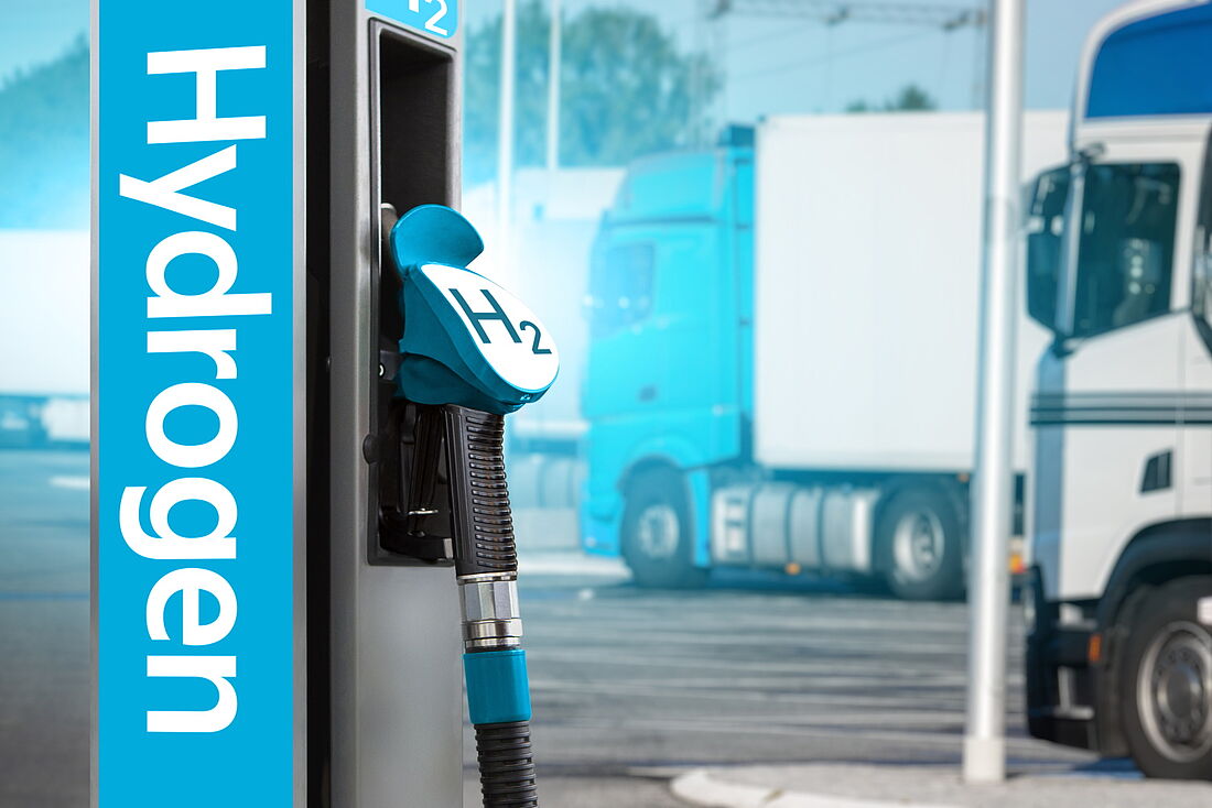 Im Vordergrund ist eine Tanksäule einer Wasserstofftankstelle zu sehen, im Hintergrund parken LKW. 