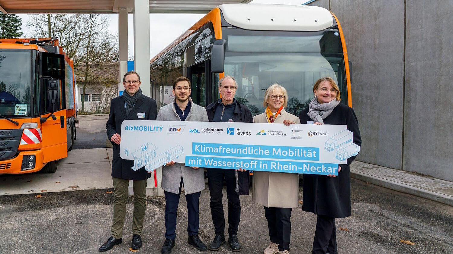 Fünf Verantwortliche stehen mit einem Plakakat vor einem Bus und einem Abfallsammelfahrzeug an der neuen Wasserstkfftankstelle.